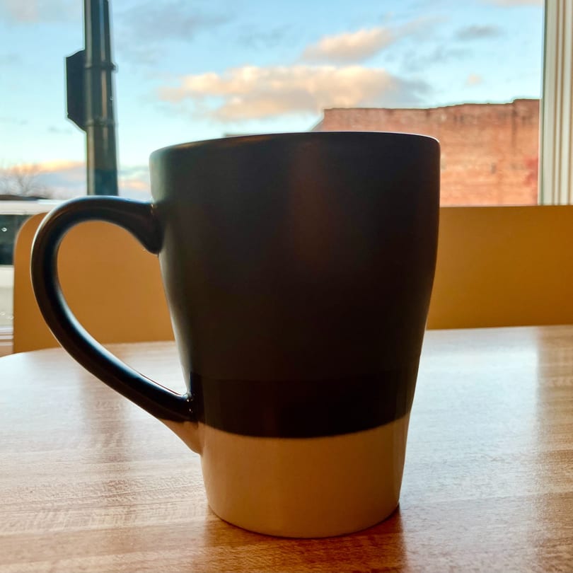 A two-toned coffee mug sits on a wooden table with a window view of the sky.