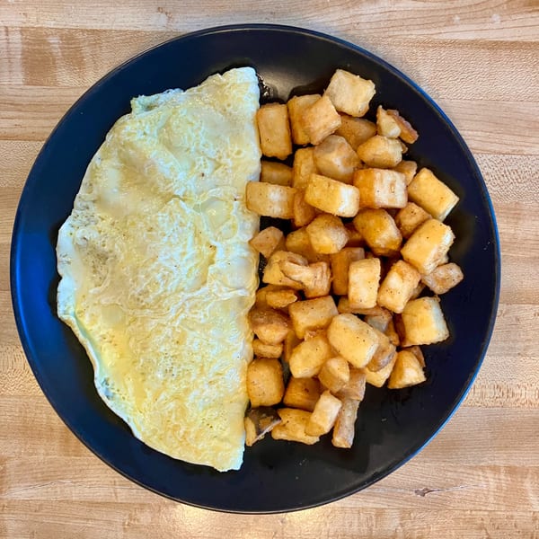 A black plate holds a fluffy omelet and a pile of crispy home fries on a wooden table.