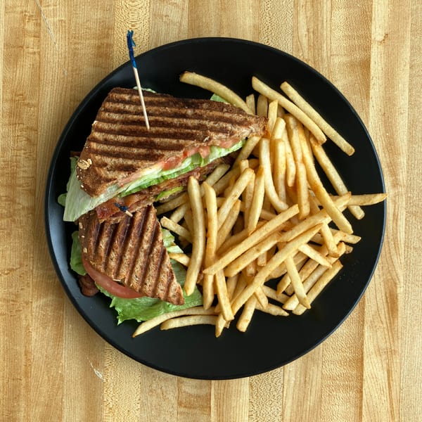 Grilled sandwich with lettuce, tomato, and bacon, served with a side of french fries on a black plate.
