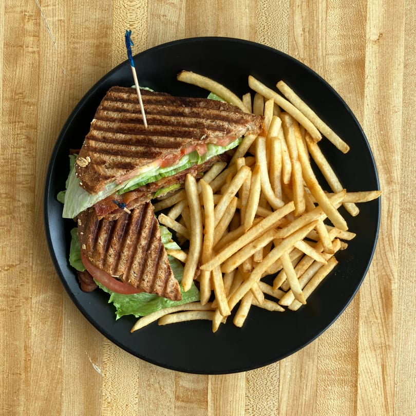 Grilled sandwich with lettuce, tomato, and bacon, served with a side of french fries on a black plate.