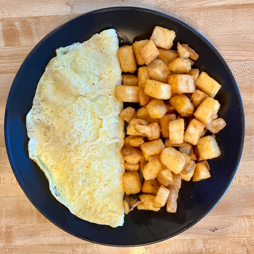 A black plate holds a fluffy omelet and a pile of crispy home fries on a wooden table.