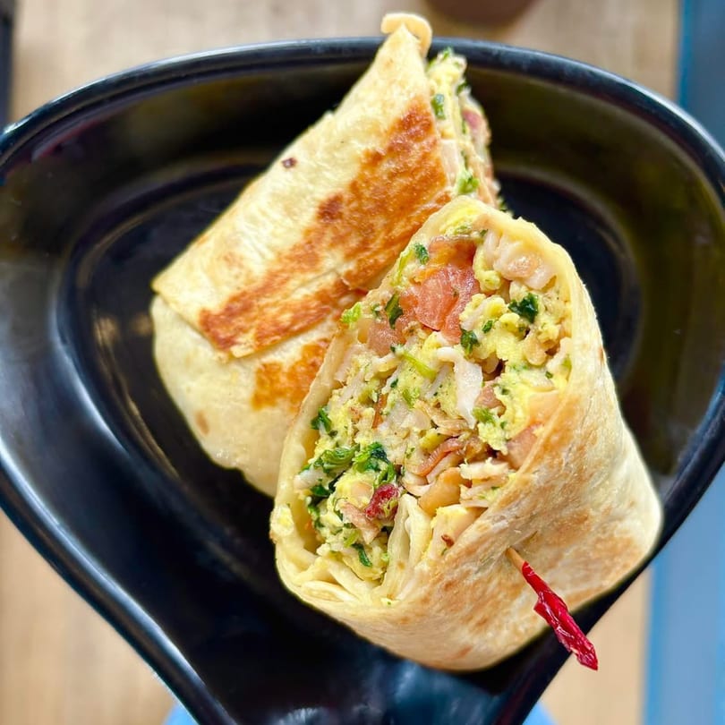 Two breakfast burritos, one cut open to show scrambled eggs, bacon, and greens, on a black plate.