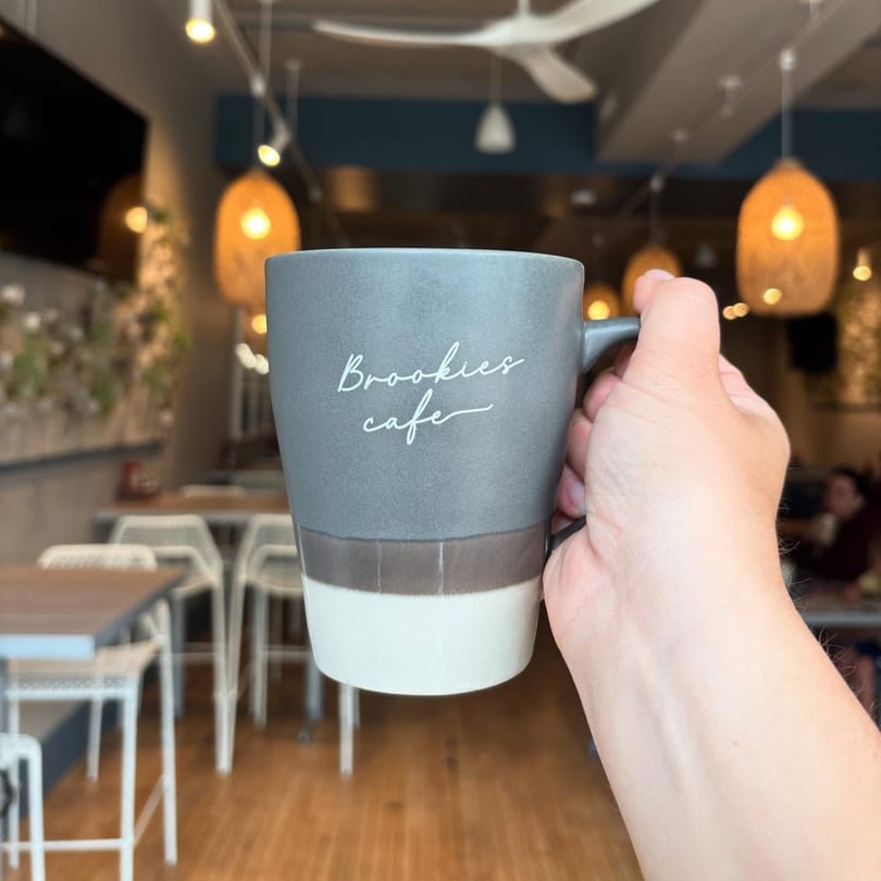 Hand holding a Brookies Cafe mug with the restaurant interior and warm lighting in the background.