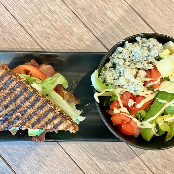 A grilled sandwich with bacon, lettuce, and tomato next to a salad with blue cheese, avocado, and tomatoes.