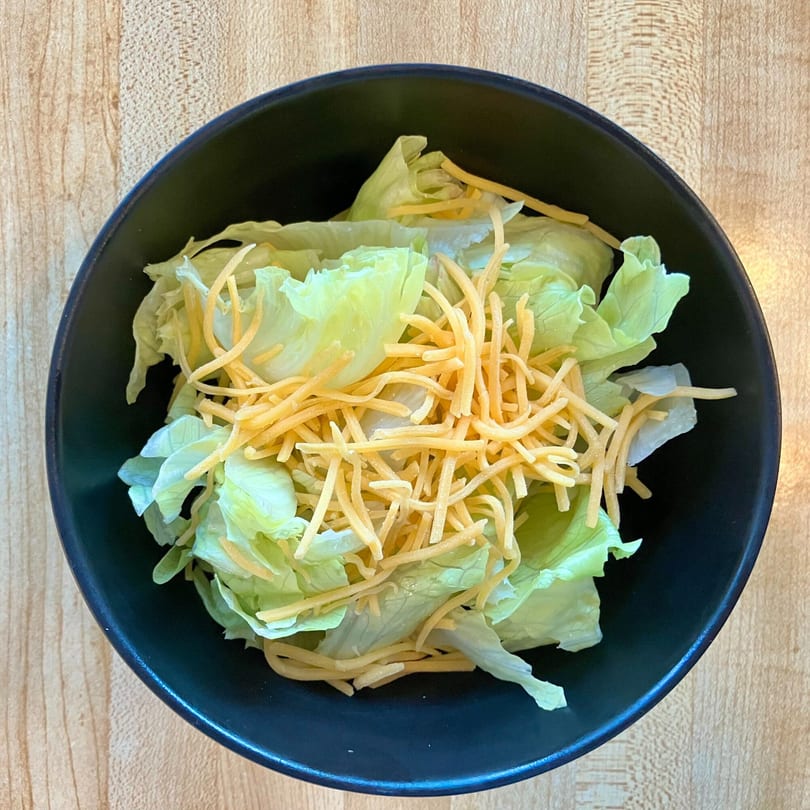 Overhead view of a black bowl filled with green lettuce and shredded cheddar cheese.