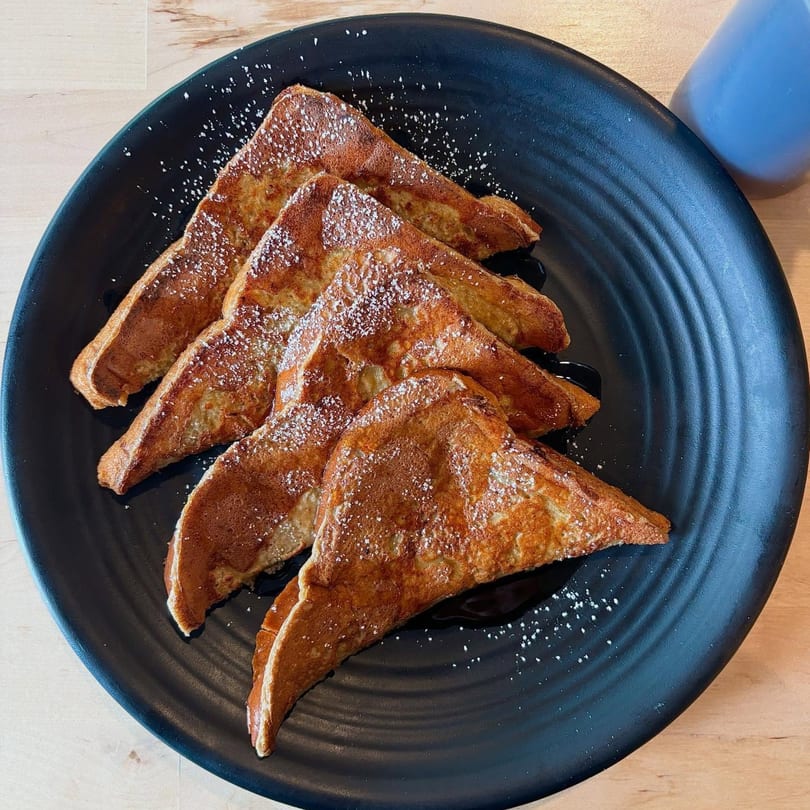 Four triangular pieces of golden French toast, drizzled with syrup and powdered sugar, on a black plate.