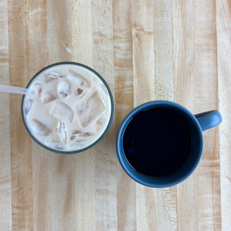 Overhead view of an iced coffee with a straw and a dark coffee in a blue mug on a wooden table.